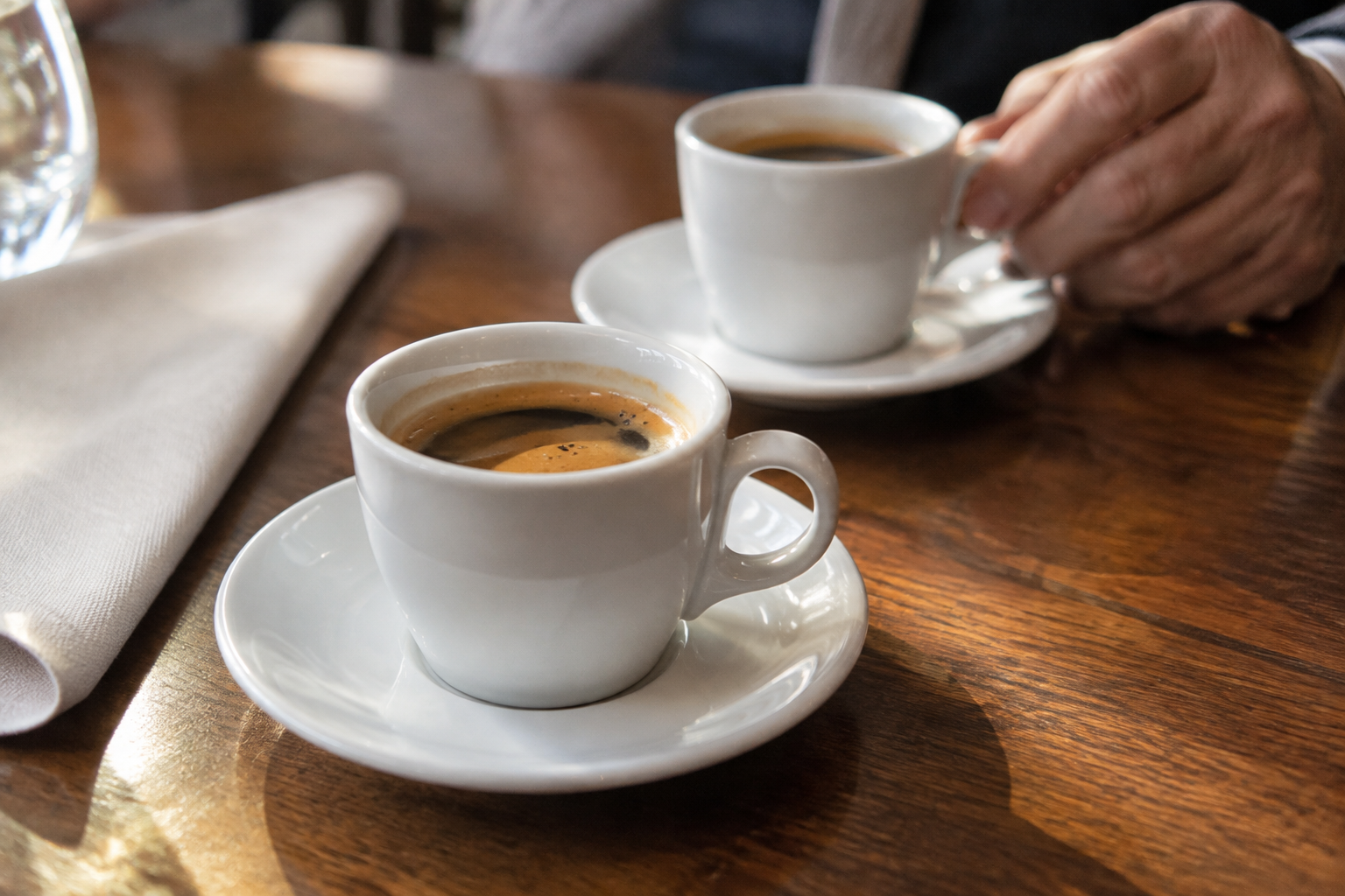 Deux tasses blanches de café sur leurs soucoupes, posées sur une table en bois, avec une main tenant l’anse de la tasse à l’arrière-plan.